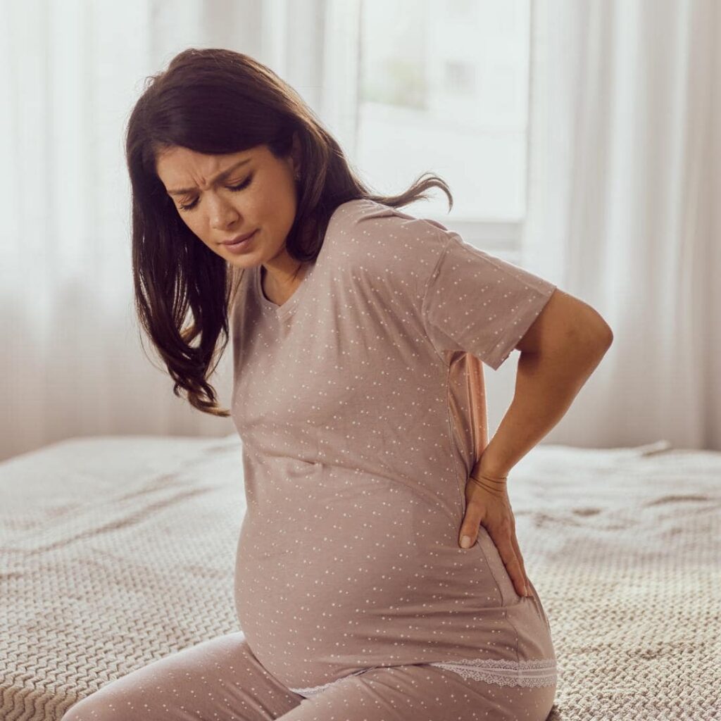 photo of pregnant person sitting in their bed holding their back due to back pain