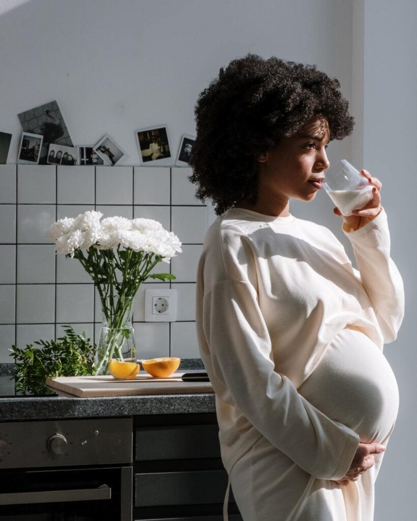 Photo of black pregnant person, with curly hair drinking a glass of water standing in front of kitchen counter with white flowers. 
