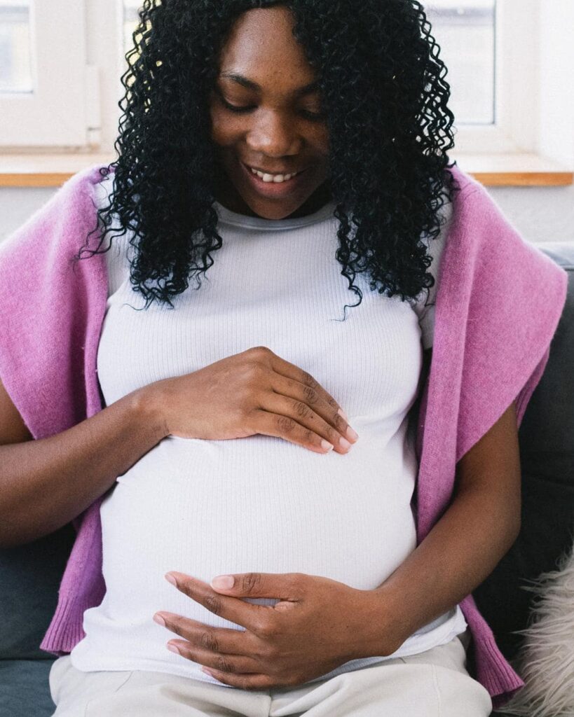 Photo of pregnant black woman wearing a white shirt with pick sweater over her shoulders, sitting on a couch. 