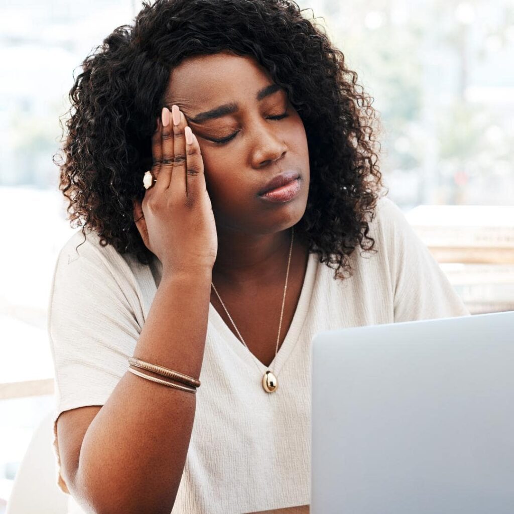 Photo of black woman sitting at her desk with her eyes closed and beginning to have  overload burnout. 