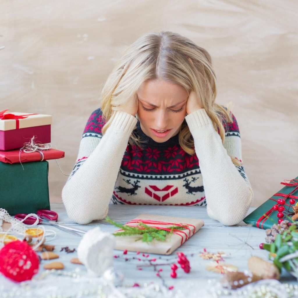 Photo of blonde hair woman in holiday sweater, sitting at a table surrounded by holiday wrapping.  She's holding the sides of her head in frustration. 