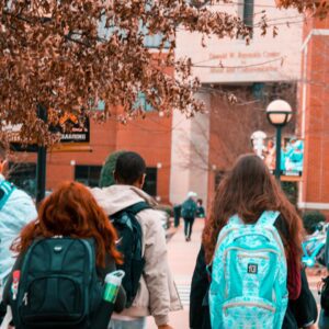 Photo of teens walking into a high school.  They are all wearing backpacks. 